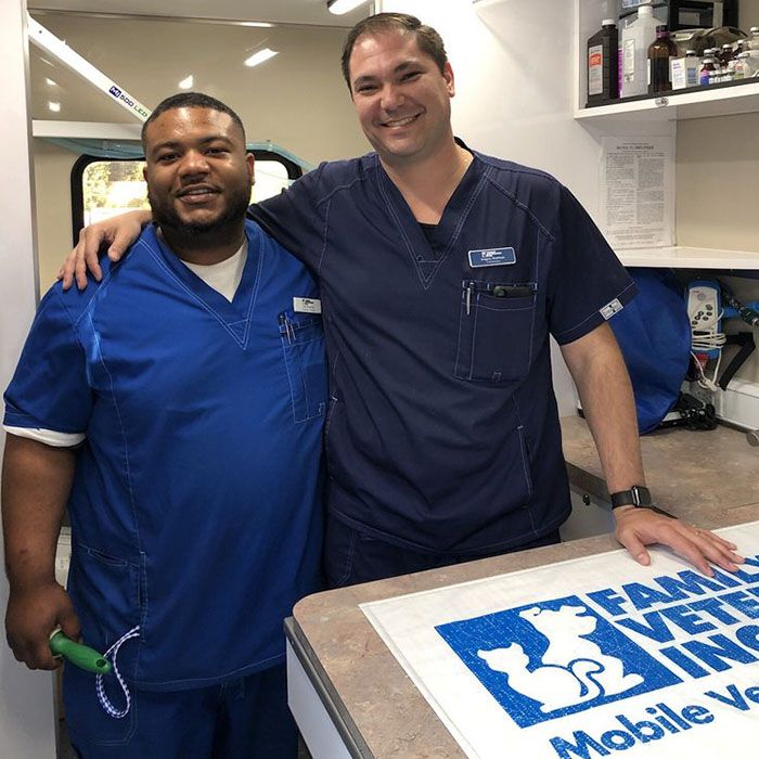 Two men wearing blue scrubs stand smiling in a mobile veterinary clinic, with one holding a toy.